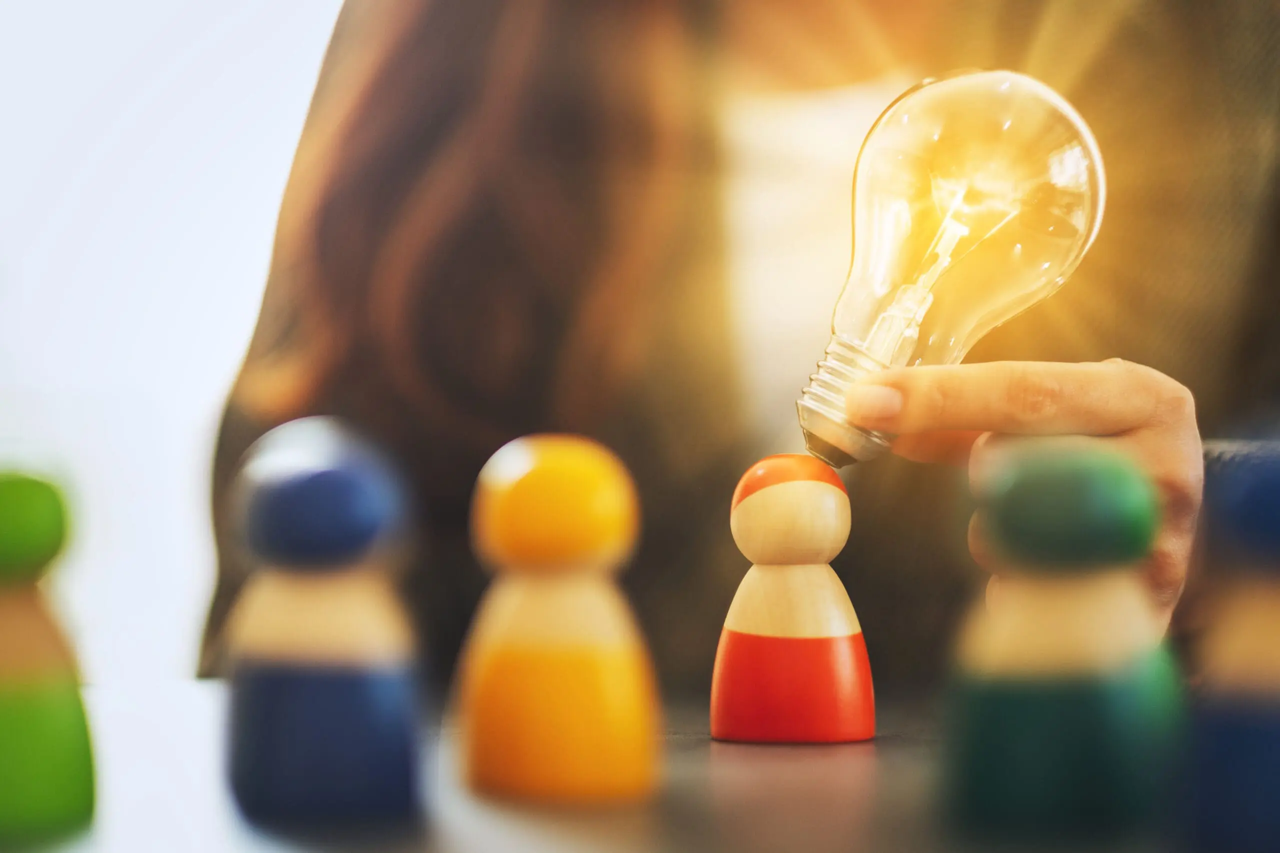 Mano sosteniendo un foco de luz brillante e iluminado sobre una figura de madera específica destacada entre un grupo. Representa la selección de innovación y del top 5% del talento tecnológico de élite (Hub en Honduras), aportando ejecución impecable para el mercado de Austin y Centroamérica.