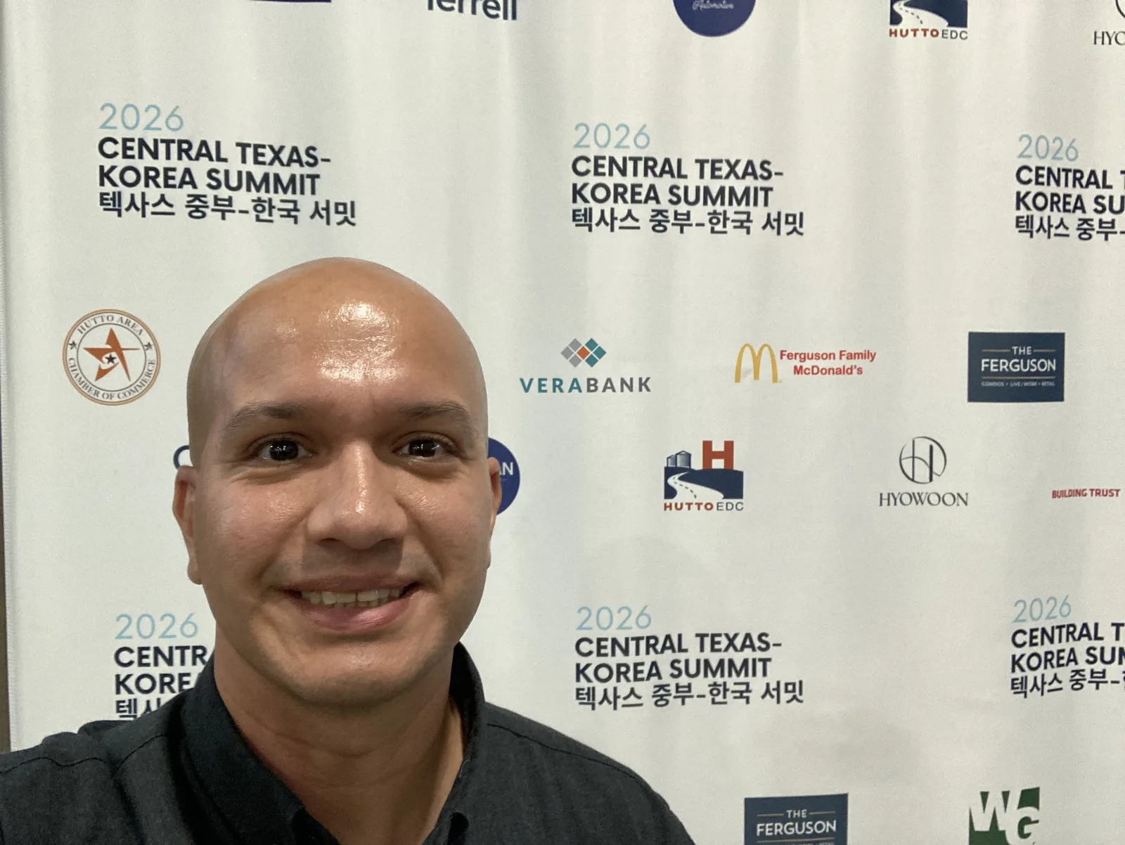 Integrante del equipo de Andes Development frente al backdrop oficial de la Cumbre Central Texas-Corea 2026 en Taylor, Texas, con los logotipos de los patrocinadores VeraBank, Hutto EDC, Hutto Area Chamber of Commerce y The Ferguson, y el nombre del evento en inglés y coreano.