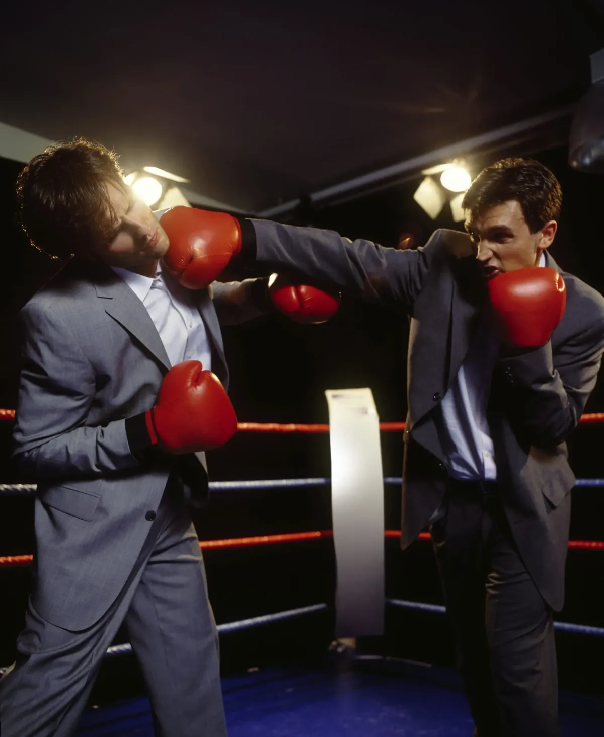 Two businessmen in gray suits and red boxing gloves fighting in a ring. A metaphor for the traditional conflict and debate over which programming language to choose, a problem solved by the Dual Stack approach (Python and Laravel).