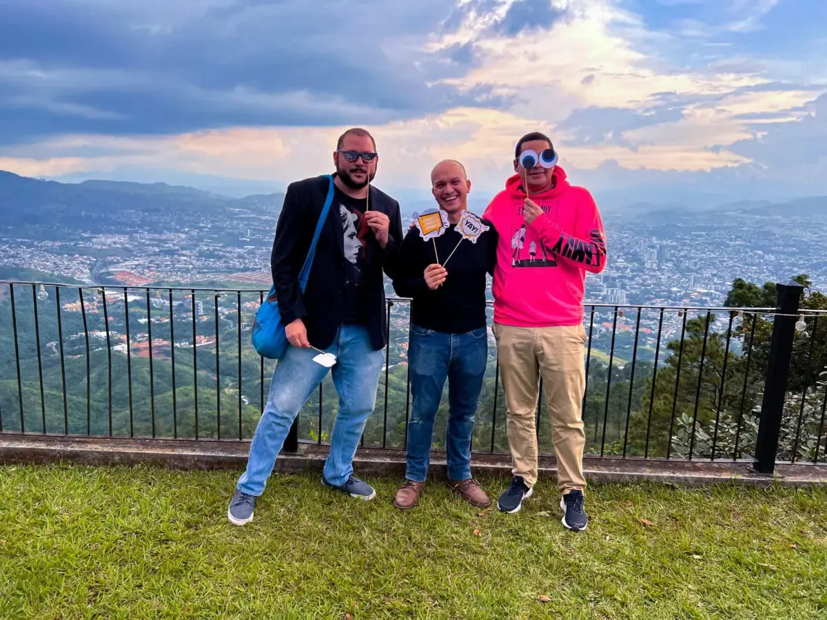 Three Andes Development team members sharing a fun moment with festive props during the company's 12-year celebration. The outdoor photo shows a spectacular view of the city and mountains in the background, reflecting the camaraderie at the agency.