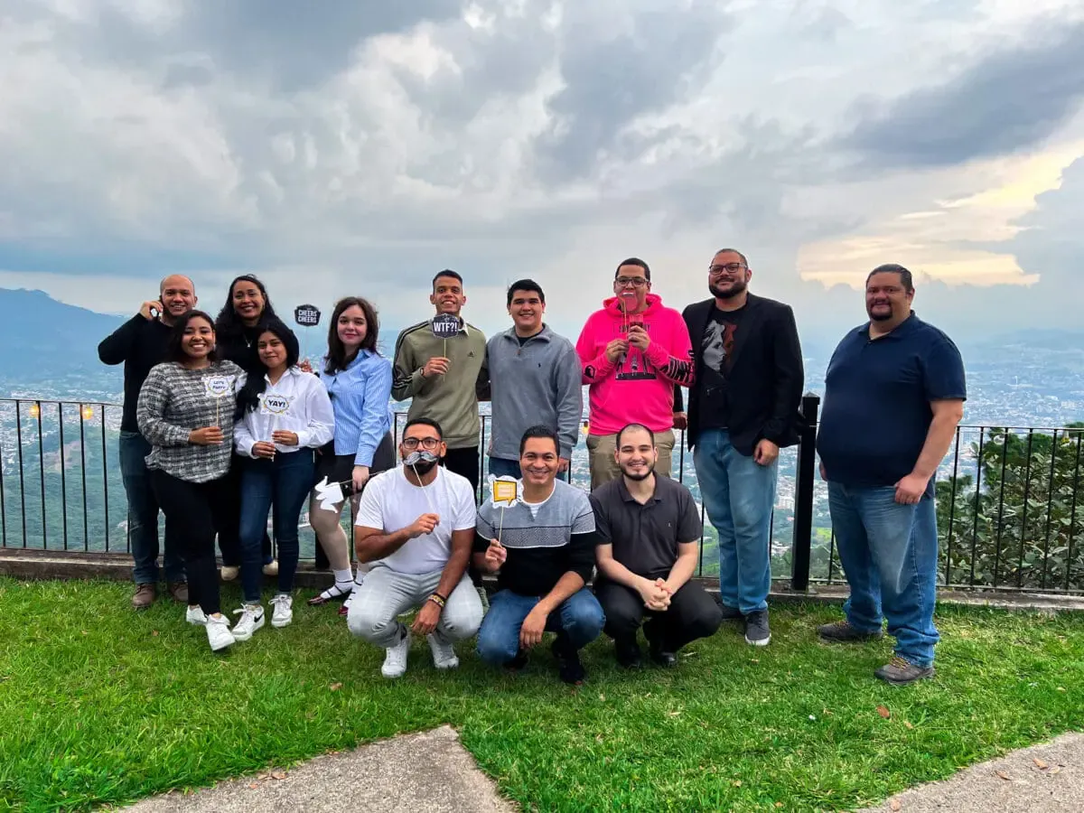 Group photo of Andes Development team members posing with creative photo booth props at a scenic lookout. It captures the 12th anniversary celebration and the company's excellent work environment with the city in the background.