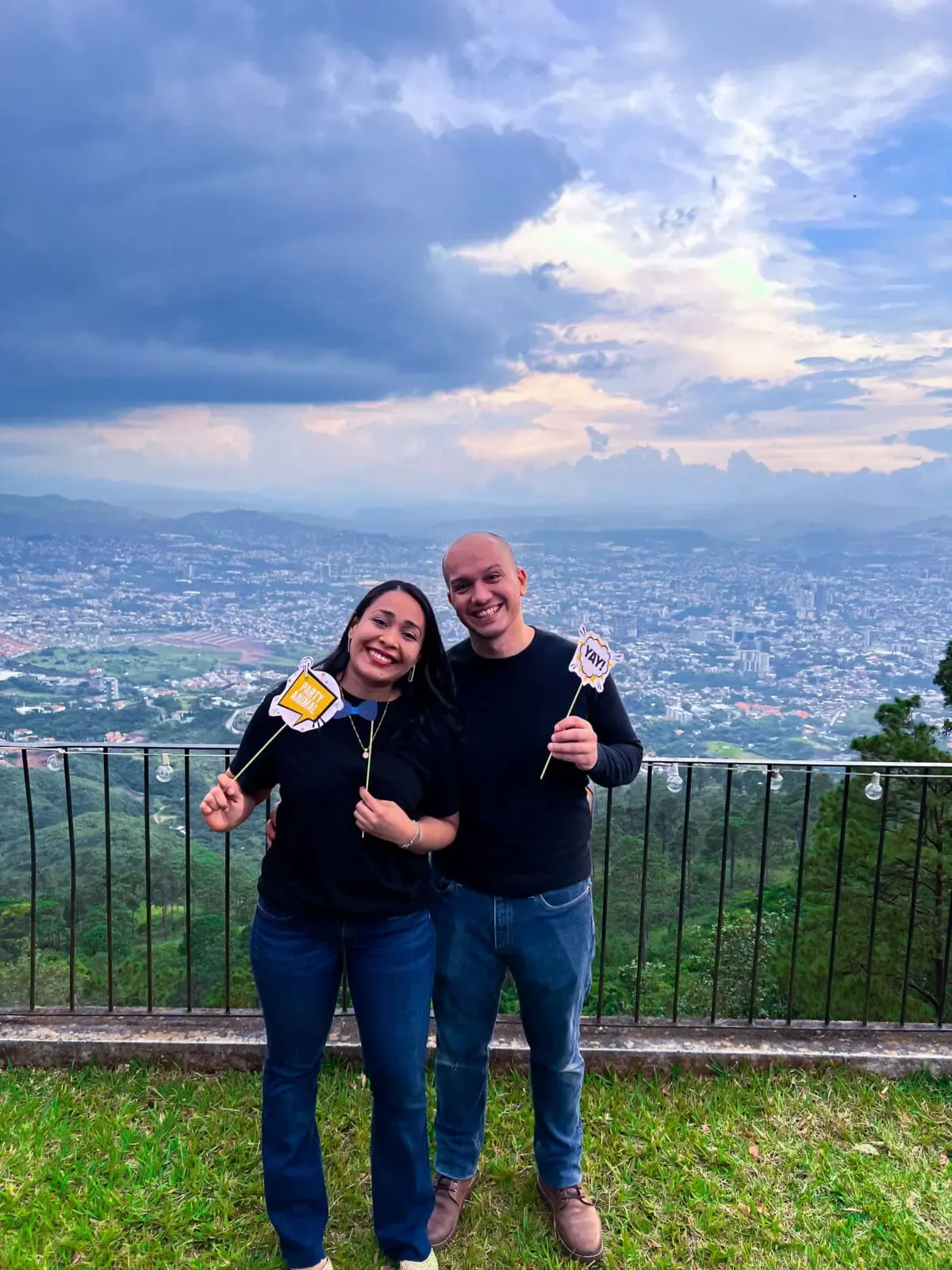 Dos miembros del equipo de Andes Development sonriendo y posando con divertidos accesorios de cabina de fotos (como un letrero de 'Party Animal') durante la celebración del 12º aniversario. De fondo se aprecia una hermosa vista panorámica de la ciudad al atardecer.